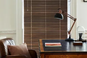 Home office with cream walls, traditional wooden desk, leather tub chair, black anglepoise lamp and tall narrow window dressed in dark wooden venetian blind, lowered, louvres almost totally closed.