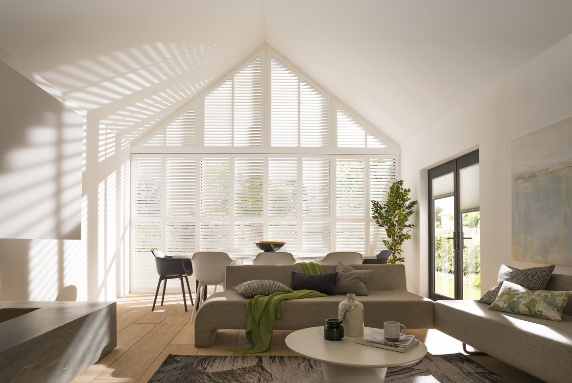 Large open-plan living-diner with gable end windows and doors dressed in white shutters, feature fireplace on left, beige sofa opposite with dining table and chairs in front of gable end at the back.