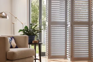 Cream living room with brown soft chair, wooden side table, two skylights and bifold doors dressed in tracked door shutters, partially open.