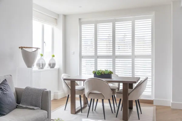 full height white shutters in white room with grey sofa and small dining table with four grey chairs