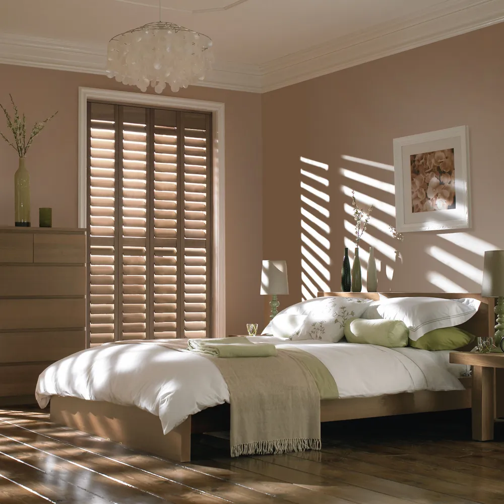 Bedroom with pale mink walls, mid wood floor and furniture, white beeding  and two tall windows dressed in mid brown wooden shutters, closed with louvres open and sun rays casting on bed, wall and floors.