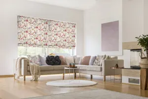 Neutral lounge with L shaped sofa with accent cushions in soft pink and floral matching two Roman blinds at window behind with medium sized pink and beige flowers, accent grey knot-shaped cushion on sofa.
