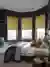 Large bedroom with bay window with three ochre faux velvet Roman blinds, wooden floor, navy walls, bed with white bedding and grey quilted throw, modern pendant light.