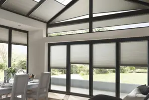 Open plan lounge diner glass gable end extension with white wood table and chairs, sofa with throws, and grey Duette blinds to doors and windows and angled roof windows.