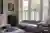 Cream lounge with bay window dressed in white birch cafe style shutters, closed with louvres open, silver grey sofa and large footstool as table, wooden side table and dark wood apothecary cabinet.