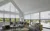 Living room with gable end windows to left and run of windows and doors on adjacent wall, all dressed in soft white duette blinds, lowered halfway, pale grey carpet, mid grey L-shaped sofa and footstools with cushions and throws in olive, dark grey and dusky orange.