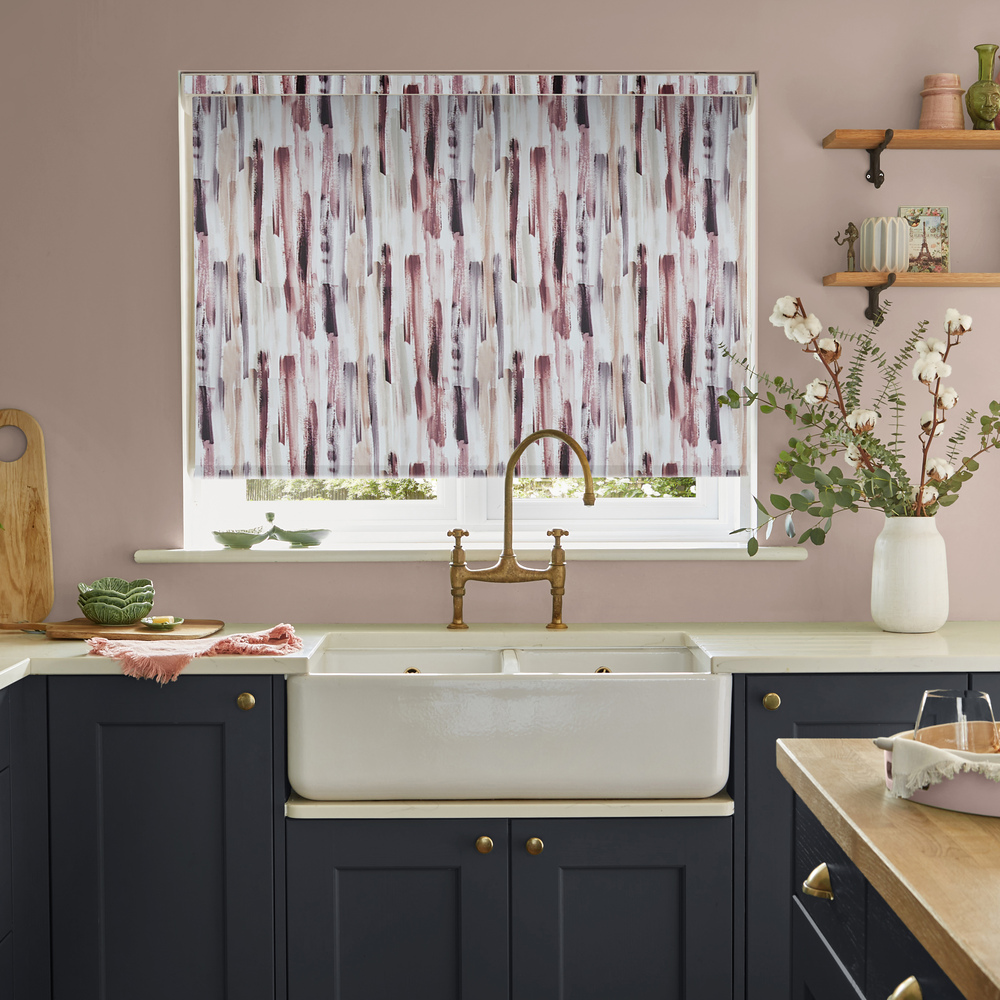 Kitchen with a window dressed with a roller blind in a pink, grey and beige waterfall pattern, dark blue cabinets, dusky pink walls and behind a butlers sink with antique brass mixer taps.
