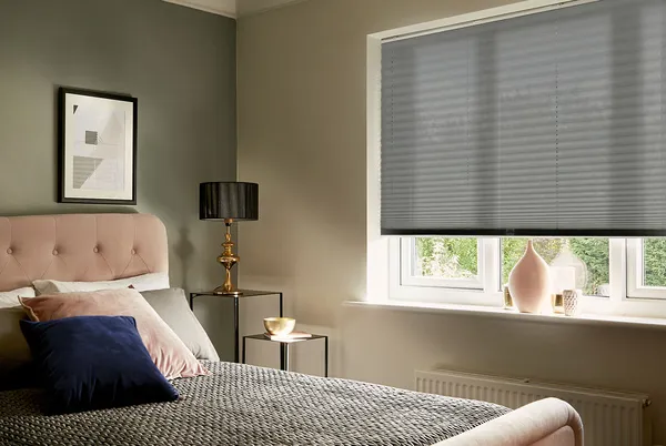Tight shot of bedroom with one dark and one light brown wall, mid grey pleated blind three quarter lowered at window, bed with dusky pink head and foot, grey bedding, pink, brown and grey accessories.
