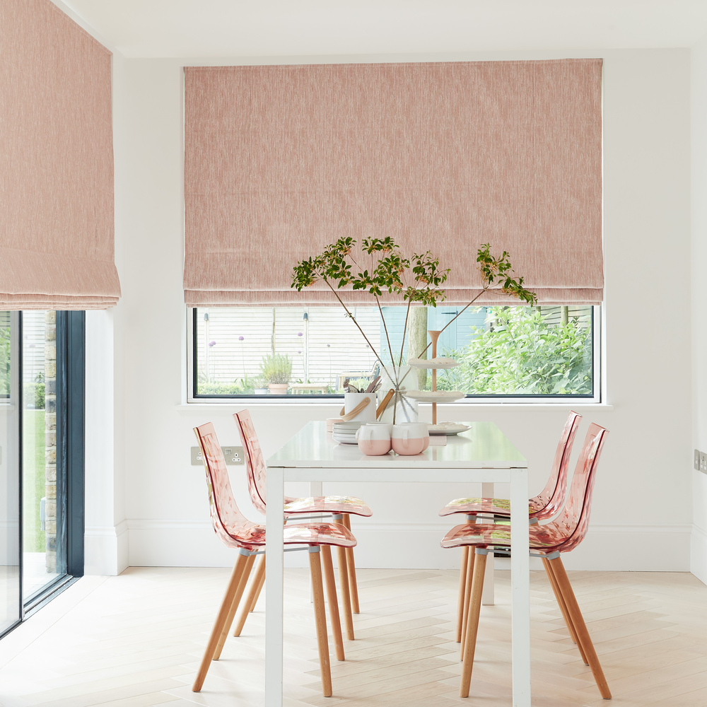 Dining room in white with window to rear and patio doors to left dressed in Roman blinds with orange-red geometric linear design on white background, with modern white table and pink marl chairs with pink and white crockery and vase with tall stemmed greenery.