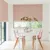 Dining room in white with window to rear and patio doors to left dressed in Roman blinds with orange-red geometric linear design on white background, with modern white table and pink marl chairs with pink and white crockery and vase with tall stemmed greenery.