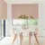 Dining room in white with window to rear and patio doors to left dressed in Roman blinds with orange-red geometric linear design on white background, with modern white table and pink marl chairs with pink and white crockery and vase with tall stemmed greenery.