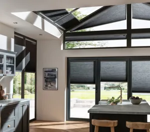 Gable end kitchen with white walls, doors and windows dressed in slate duette blinds at varying open heights, dark grey kitchen units, island with grey marble top with sink, wooden chairs, light rays on walls and floor.