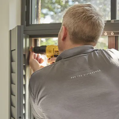 Installer using electric screwdriver to install grey shutters at a window.
