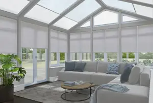 Rectangular conservatory with dark wood floor, pale grey L-shaped sofa, large rug and leafy plant in pot by door with pale lilac roller blinds on side windows and off-white pleated blinds in roof.