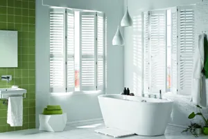 White bathroom with one walled tiled in olive green with window either side of corner dressed in white shutters, partially open above a bath.