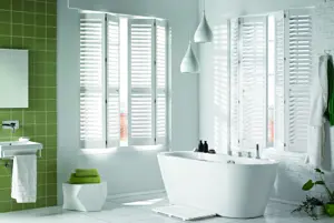 White bathroom with one walled tiled in olive green with window either side of corner dressed in white shutters, partially open above a bath.