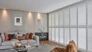 White wooden shutters fitted to a wide window in a living room with a sofa, chair and coffee table