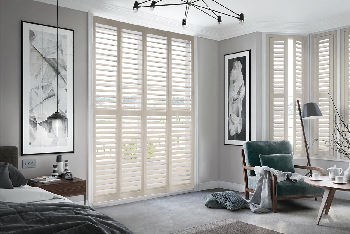In a bedroom that has a bed, sidetable, armchair and coffee table are Vintage Beige Shutters. The Yellow coloured Shutters are fitted to a series of tall windows in the bedroom which is decorated in neutral grey tones.