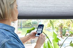 A woman looks at an app on her smartphone to control the duette blind she's standing next to.