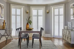 Floor to ceiling height white shutters fitted to three windows in a dining room with a dark wooden dining table and chairs