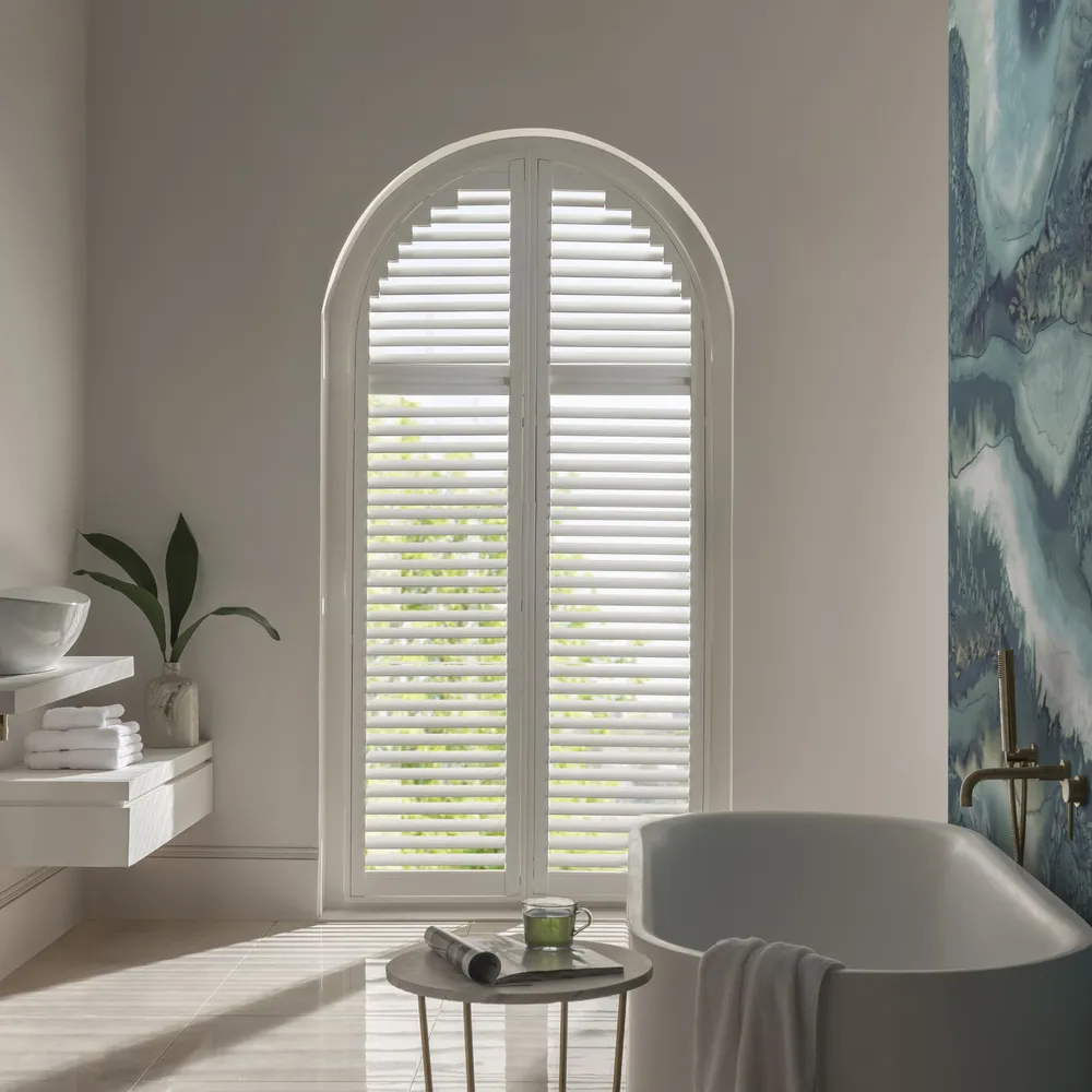 White bathroom with large arch window dressed in white shutters, closed with louvres open, abstract blue feature wall on right beside bath.