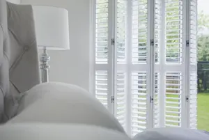 Close in of a pale grey bedroom with grey bed and white tracked shutters folded partially back, louvres open.