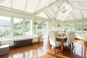 Large conservatory with off-white shaped roof blinds, dining table and chairs in centre on highly polished wooden floor flooded with sunlight