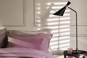 Close up in bedroom with cream panelled walls, bed with dusky pink bedding and indigo throw, small tall dark wood bedside table with dark anglepoise lamp and rays of light created by shutter louvres on the wall behind.