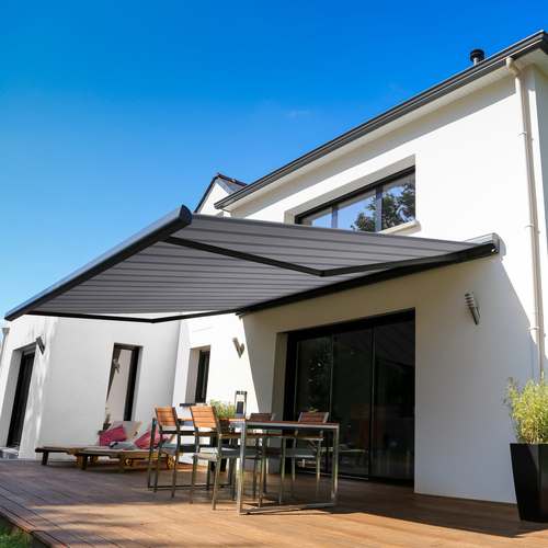 Exterior of a large modern white-rendered house with a wooden deck, contemporary table and chairs underneath an extended grey wide-striped awning with black metalwork.