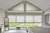 Bedroom in pale greys with beige Duette blinds at the floor to ceiling windows and doors, three quarters lowered and gable end windows, lowered, bed with grey headboard, white bedding with grey woolen throw and low bench in front of windows.