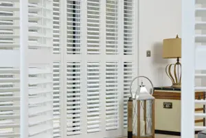 Close in through open tracked door shutters on a room with a gable end window, also dressed in white tracked shutters, closed with louvres open.