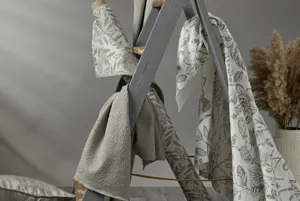 Still life shot of a small grey wood stepladder with bolts of fabric in shades of grey, patterned and plain, draped over the rungs and grey cushions and tiles around on the floor.