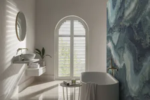 White bathroom with bath on right against feature wall with abstract marble-effect in blues and arched window to rear dressed in white shutters, louvres open.