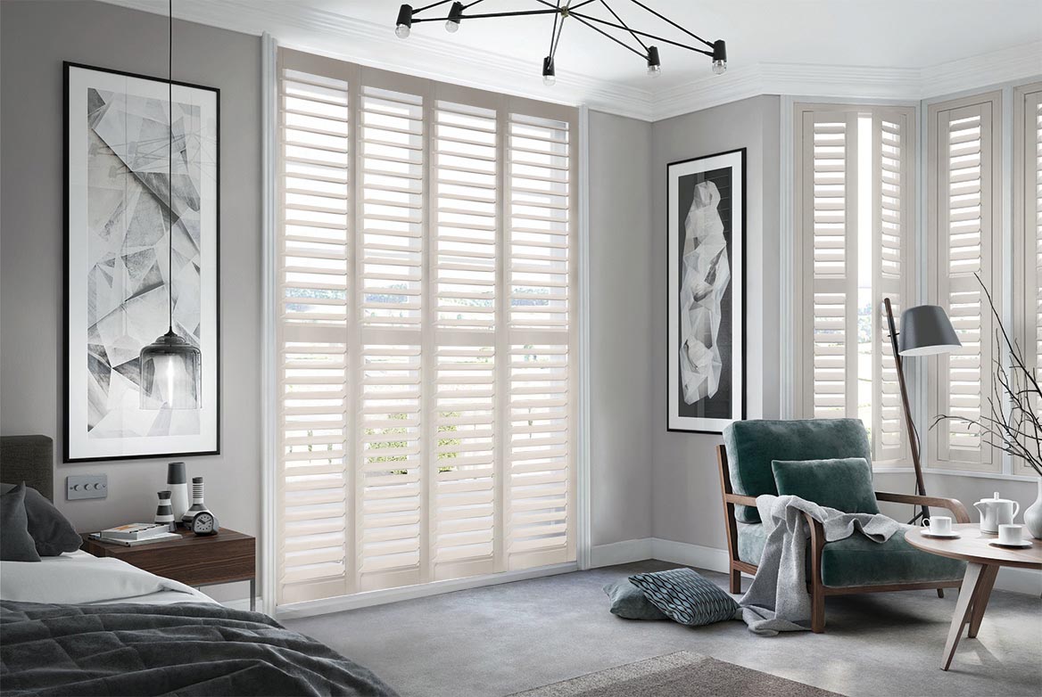 In a bedroom that has a bed, sidetable, armchair and coffee table are Country Meadow Shutters. The Ivory coloured Shutters are fitted to a series of tall windows in the bedroom which is decorated in neutral grey tones.