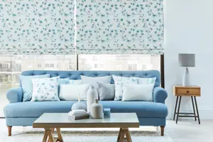 Close shot of lounge with pale blue walls, grey flooring with pale blue rug under low trestle table with blue accessories, blue sofa and cushions in front of large windows dressed in two Roman blinds of sprigs of blue flowers on white background.