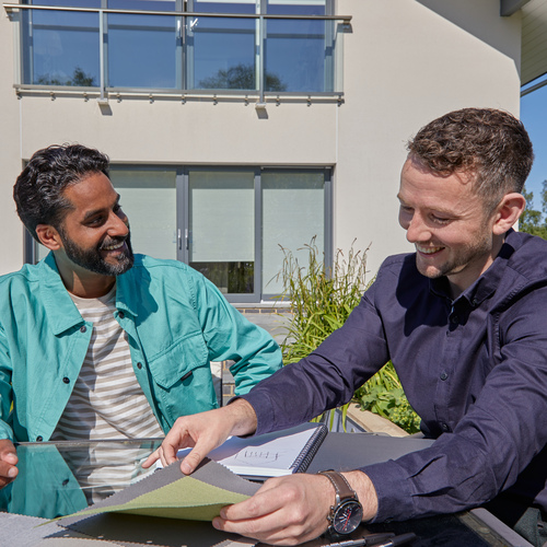 Exterior of modern house, two men seated at a table discussing awning samples spread out on the table.