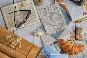 Moodboard of summer vibes with fabric and shutter samples in sea blue, cream and natural wood, seascape postcards, shells, croissant, coffee and sunglasses on an open book.