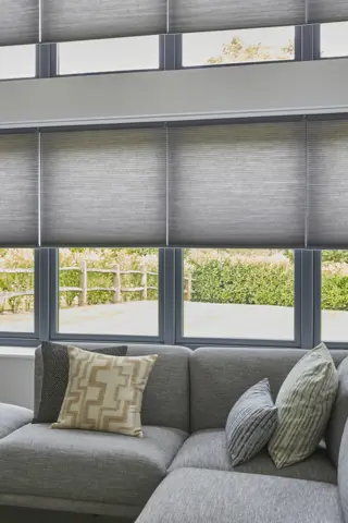 Pale grey living room with to sets of four windows one above the other dressed in pale grey duette blinds lowered halfway, L-shaped grey sofa with cushions in chartreuse and blue patterns.