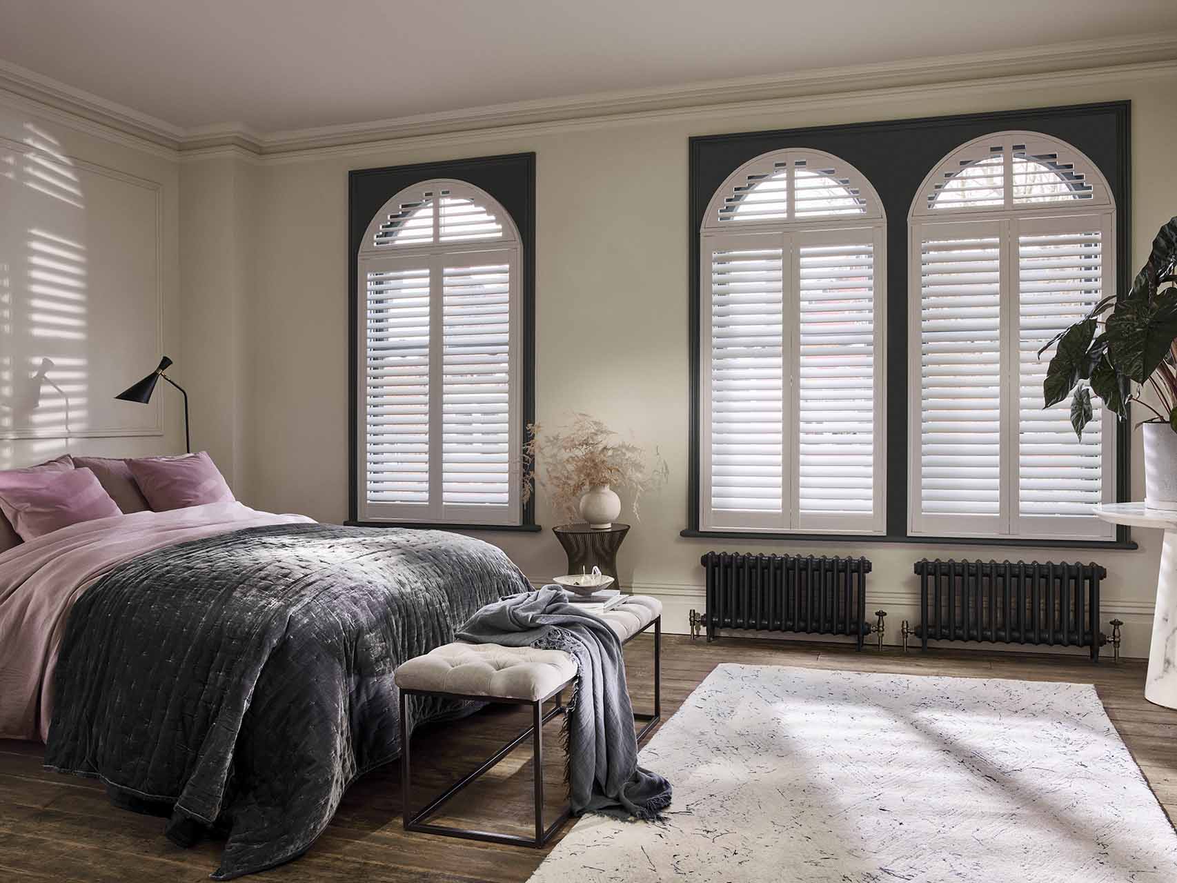 Bedroom with three windows in an arch shape with dark blue trim, dressed in white arched shutters, louvres open, room in soft white with dark wooden floor, bed dressed in usky rose pink bedding with grey velour throw, cream upholstered metal stool at bed end with grey wool throw, white rug with dark speckling, marble pedestal table with large leafy plant and .