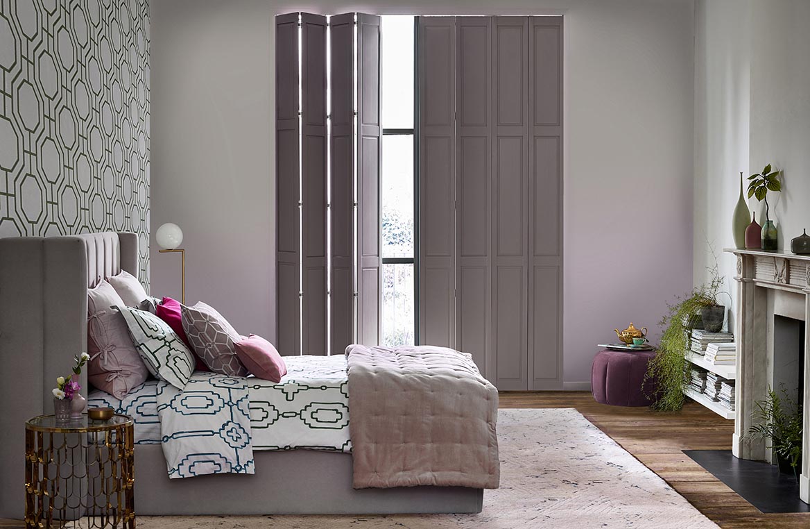 Mauve coloured solid tracked shutters fitted to a tall rectangular window in a bedroom decorative in repeating geometric designs 