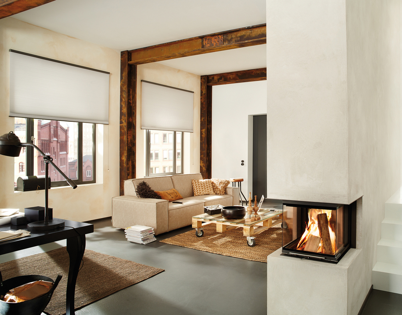 Loft apartment style  lounge room with beams and feature pillar fire, lit. and two large windows dressed in pale Duette blinds slightly lowered,