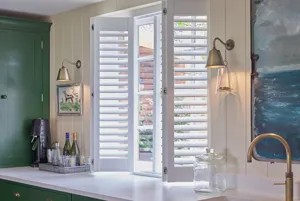 Close up on kitchen with cream wooden walls and green units with white tops and window with white shutters partially folded back, louvres open.