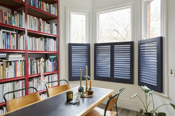 Dark blue cafe shutters fitted to a bay shaped window in a room with a dining table and book shelf 