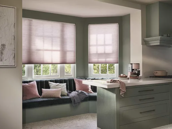 Contemporary kitchen with banquette seating under bay windows dressed in soft pink Duette® blinds drawn halfway, co-ordinating cushions on blue seating, kitchen counters in soft grey with white top.