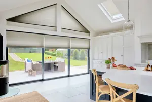 White gable end kitchen diner with white tiled floor, white units to the side, island dining area with blue units and white worktop and skylight, gable end windows and doors all dressed in mid grey duette blinds, skylight blind three quarters lowered, gable shape fully lowered and door blinds partially lowered.