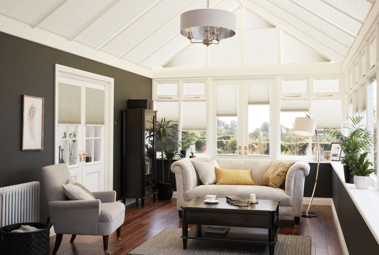 Conservatory with dark brown walls, polished dark wood floor with brown rug under dark wood coffee table, pale greige sofa and chair, with side and roof windows dressed in cream duette blinds at varying heights.