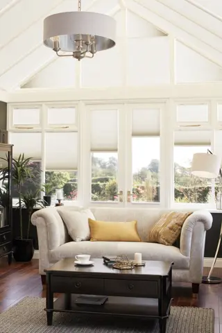 Conservatory with dark brown walls, polished dark wood floor with brown rug under dark wood coffee table, pale greige sofa and chair, with side and roof windows dressed in cream duette blinds at varying heights.
