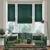 Pale grey lounge with bay window dressed in two narrow and one wide Roman blinds in deep green with pale wood floor and large faded geometric patterned forest green rug with table and green accessores, green sofa with cushions in dark green and cream.