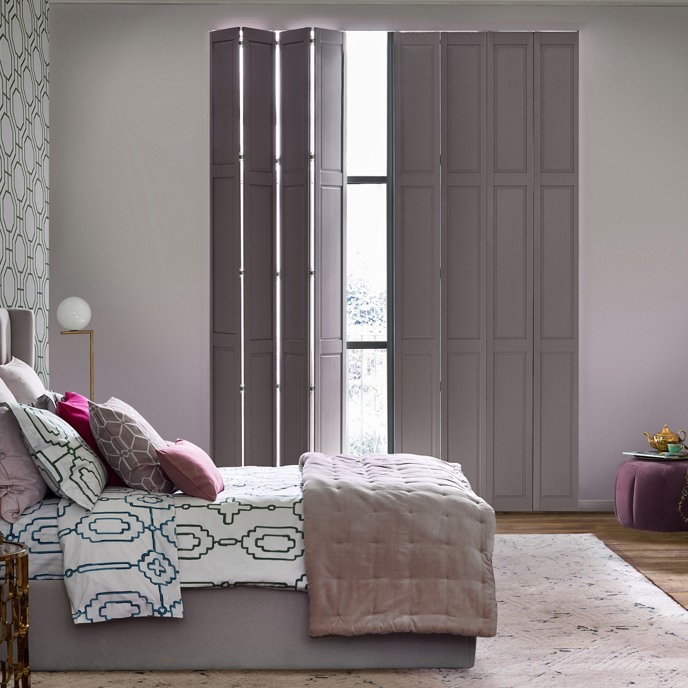 Solid tracked shutters in mauve, one partially open in a bedroom with pale mauve wall, geometric pattern wallpaper to left with mauve bed dressed in geometric bedspread with mauve throw, dark wood floor.