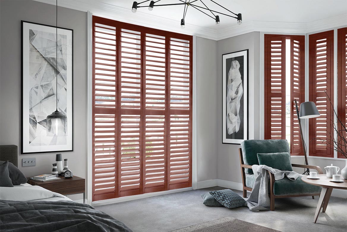Burley Orchard Red Shutters have been fitted to tall windows in a bedroom. The Shutters are Natural, Brown coloured while the bedroom is decorated with grey tones.